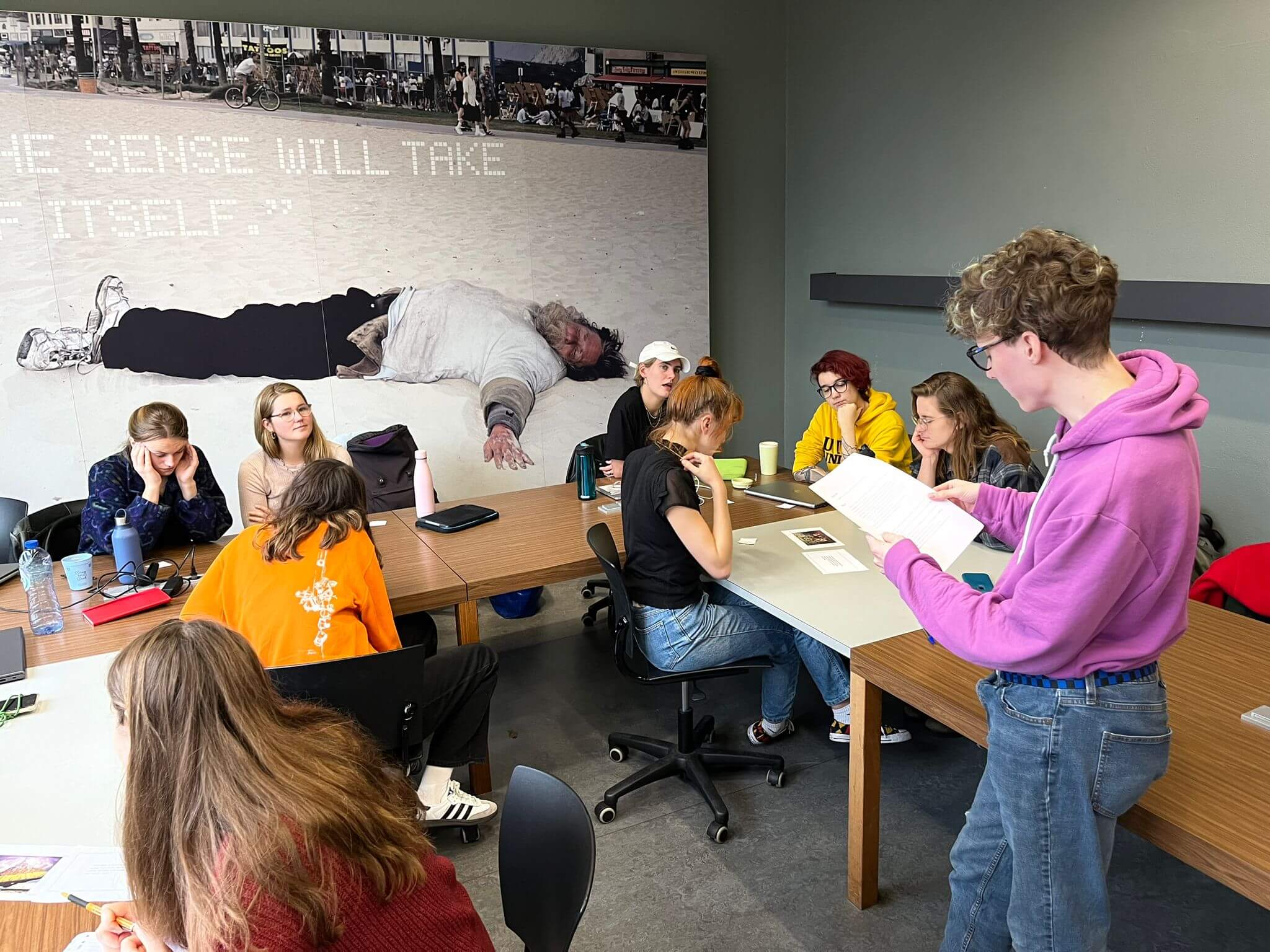Photo of me in a purple hoodie explaining playtest rules for a tabletop game to around 10 university students in groups